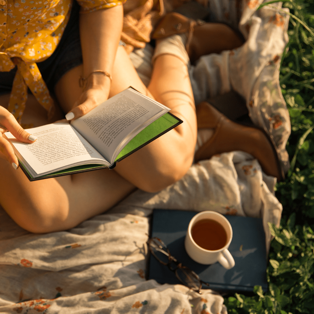 Woman reading a book from morning basket on blanket on grass on a sunny day with cup of tea